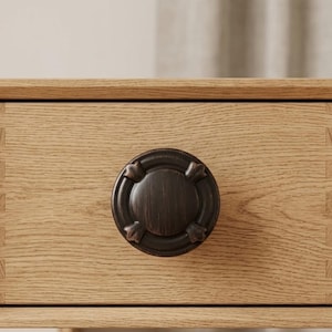 May include: Close-up of a wooden drawer with a dark bronze-colored round knob. The knob has a circular design with four decorative elements. The drawer is made of light-colored wood with visible grain and dovetail joints.
