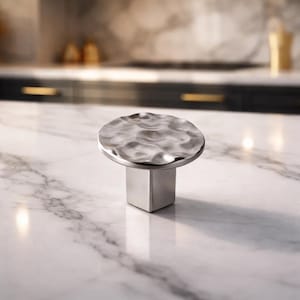May include: A silver cabinet knob with a hammered, textured surface. The knob has a round top and a square base. The hardware is made of metal and is designed for use on cabinets or drawers.