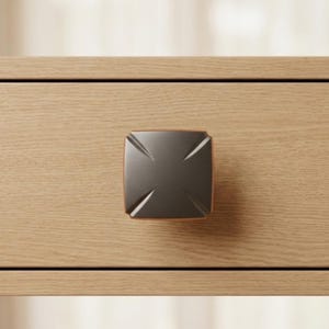 May include: A square, dark bronze cabinet knob with a cross-shaped design. The knob is mounted on a light-colored wooden drawer. The hardware has a modern, geometric aesthetic.
