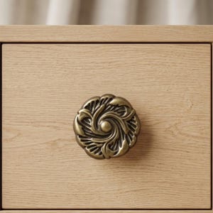 May include: Close-up of a decorative brass drawer pull with an ornate floral design. The drawer pull is mounted on a light-colored wooden drawer front, showcasing the hardware's intricate details and warm tones.