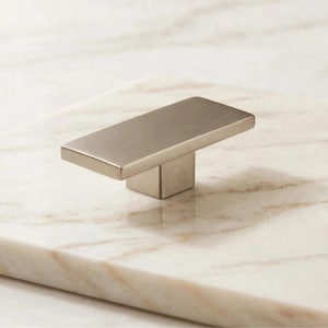 May include: A rectangular cabinet knob with a brushed metallic finish. The hardware features a flat, elongated top surface and a central square base, showcasing a smooth, uniform texture and a muted, light silver-toned appearance.