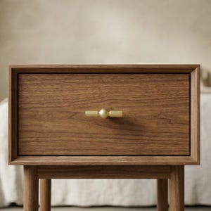 May include: A wooden nightstand with a single drawer and a gold-colored knob. The nightstand is made of light brown wood and has a simple, modern design. The drawer is centered on the front of the nightstand.