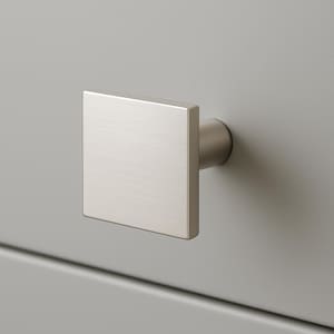 May include: A close-up of a brushed nickel square cabinet knob. The knob is mounted on a light gray cabinet door. The square knob has a modern, minimalist design and a cylindrical base.
