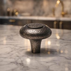 May include: A mushroom-shaped cabinet knob with a textured, hammered metal finish. The knob is a dark, muted silver color and sits on a white marble surface. The knob has a small, circular top and a flared base.