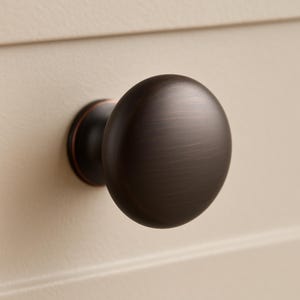 May include: A close-up of a bronze-colored cabinet knob with a rounded shape. The knob is mounted on a cream-colored cabinet door. The knob has a dark, metallic finish and a circular base.