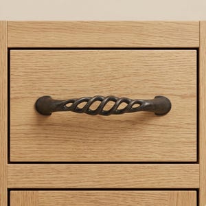 May include: Close-up of a wooden drawer with a dark bronze-colored, twisted metal handle. The drawer is made of light-colored wood with visible grain. The handle is horizontally centered on the drawer front.