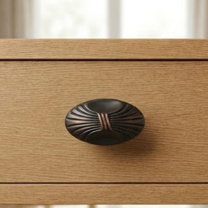 May include: Close-up of a dark bronze oval drawer pull with a fan-like design. The pull is mounted on a light-colored wooden drawer, showcasing the hardware's decorative details and contrasting colors.