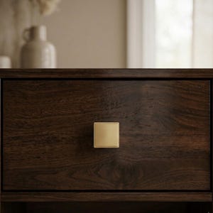 May include: Close-up of a dark brown wooden drawer with a square, gold-colored knob. The drawer's wood grain is visible, and the knob is centered on the drawer front. The background is blurred, suggesting a room setting.