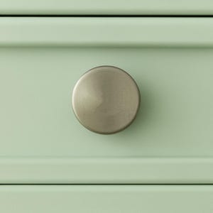 May include: Close-up of a round, brushed metal cabinet knob on a light green drawer. The knob is centered and has a slightly domed shape. The drawer has a recessed panel design.
