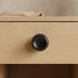 May include: Close-up of a wooden drawer with a dark, round metal knob. The drawer is made of light-colored wood with visible grain. The knob is centered on the drawer front, providing a minimalist aesthetic. The background is blurred.