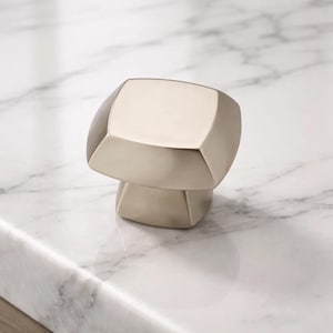 May include: A brushed nickel cabinet knob with a geometric design. The knob has a square top and a cylindrical base. The hardware is shown on a white marble surface.