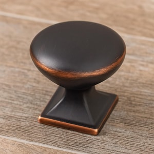 May include: A dark bronze cabinet knob with a rounded top and a square base. The knob has a copper-colored accent around the top edge and base. The hardware is designed for use on drawers and cabinets.