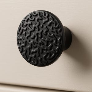 May include: A black, round cabinet knob with a textured, organic pattern on the face. The knob is mounted on a light beige surface, providing a contrast in color and texture. The knob's design adds a modern touch.