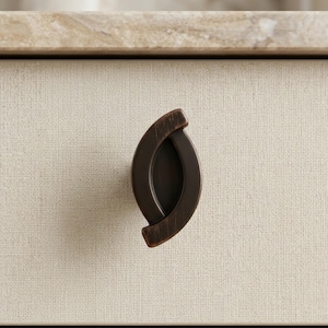 May include: A close-up of a dark bronze-colored drawer pull with an oval shape. The pull is mounted on a cream-colored, textured surface. The hardware has a modern design.