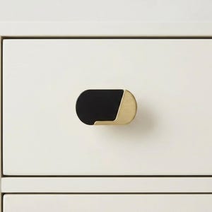 May include: A black and gold cabinet pull with a rounded rectangular shape. The pull is mounted on a white cabinet drawer. The black portion is on the left, and the gold portion is on the right.