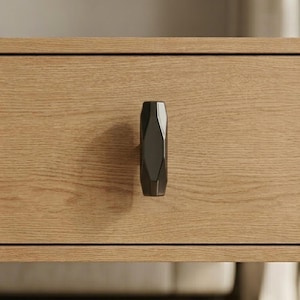 May include: A close-up of a wooden drawer with a dark gray, geometric handle. The handle has a faceted design and is vertically oriented. The wood grain of the drawer is visible, with a natural, light brown color.
