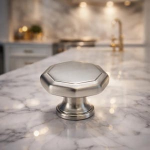 May include: A brushed nickel cabinet knob with an octagonal top and a rounded base. The hardware is set against a marble countertop, suggesting a kitchen or bathroom setting. The knob's design is modern and minimalist.