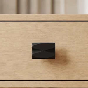 May include: Close-up of a light-colored wooden drawer with a black geometric drawer pull. The drawer pull is rectangular with a faceted design. The wood grain is visible on the drawer front.
