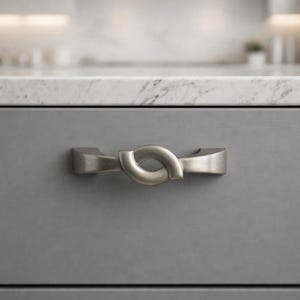 May include: A brushed nickel cabinet pull with a unique, looped design. The hardware is mounted on a gray cabinet door, with a marble countertop in the background. The pull is approximately 6 inches long.