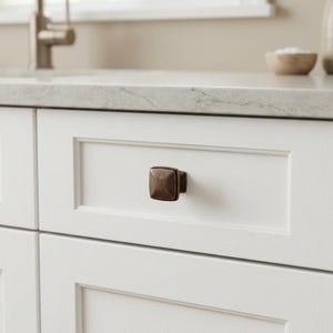 May include: A square, bronze-toned cabinet knob with a pyramid-shaped center and beveled edges, mounted on a white, flat-panel drawer front. The hardware features a dark, weathered metallic finish with visible highlights on the raised surfaces.
