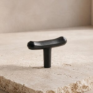 May include: A matte black T-shaped cabinet knob with a slightly curved, elongated top bar and a cylindrical base. The hardware features a smooth, solid finish and a geometric design, providing a functional grip for drawers or cabinet doors.