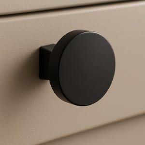 May include: A close-up of a black, round cabinet knob with a square base. The knob is mounted on a beige cabinet drawer. The knob has a matte finish and a simple, modern design.
