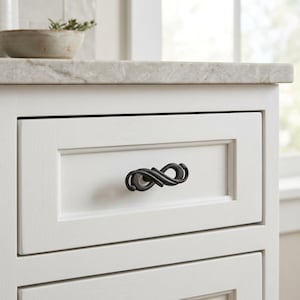 May include: A dark, oil-rubbed bronze cabinet pull with a twisted, infinity-inspired design mounted on a white wooden drawer. The hardware features a textured, metallic finish and a curved, continuous shape that spans the center of the drawer front.