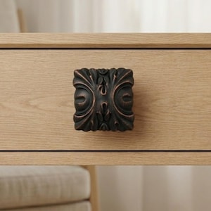 May include: A dark bronze, square cabinet knob with an ornate, symmetrical design. The knob is mounted on a light-colored wooden drawer, showcasing its decorative details and contrasting colors. The knob's dimensions are approximately 1.5 inches square.