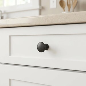 May include: A round, dark gray cabinet knob with a matte, textured finish attached to the center of a white, flat-panel drawer front. The knob features a simple, mushroom-shaped profile with a smooth, rounded top and a tapered base.