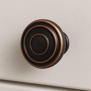 May include: Close-up of a bronze-colored cabinet knob with a circular design. The knob has concentric rings and a rounded handle. The hardware is set against a neutral, off-white background.