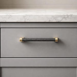 May include: A gray cabinet drawer with a black textured handle. The handle has gold-colored end caps. The drawer is part of a gray cabinet with a white countertop.