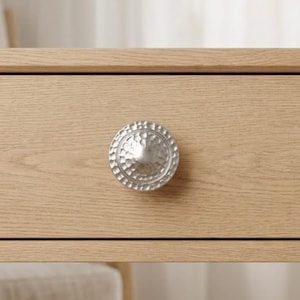 May include: A silver, hammered-metal drawer pull is centered on a light-colored wooden drawer. The round pull has a textured surface and a slightly domed center. The wood grain is visible on the drawer front.