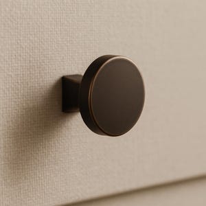 May include: A close-up of a bronze-colored cabinet knob. The knob has a circular face and a square base, mounted on a textured, off-white surface. The hardware is a simple, modern design.