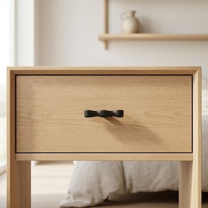 May include: A light-colored wooden nightstand with a single drawer. The drawer features a black, curved handle. The nightstand is made of light-colored wood and has a simple, modern design. The drawer is centered on the front of the nightstand.