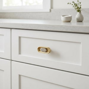 May include: A horizontal oval cabinet knob featuring a brushed gold outer frame and a smooth, matte silver center insert. The hardware is mounted on a white shaker-style drawer front, showcasing a clean, metallic finish with a dual-tone design.