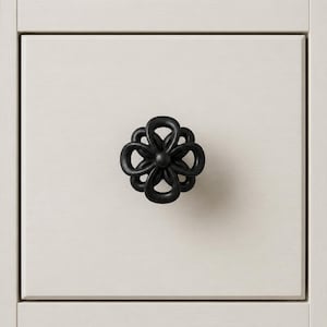 May include: A black, flower-shaped cabinet knob is centered on a white drawer front. The knob has a detailed, openwork design. The drawer front has a light wood grain texture and is part of a larger cabinet.