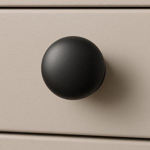 May include: A matte black, round cabinet knob. The knob is attached to a light beige drawer front. The knob is a simple, modern design, suitable for various furniture styles.