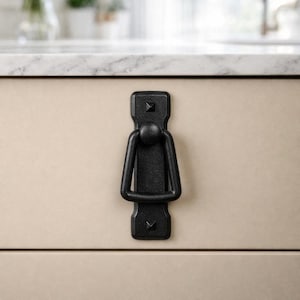 May include: Black cabinet pull with a ring handle. The rectangular backplate has a decorative, studded design. The handle is a rounded ring shape. The hardware is mounted on a light beige cabinet door.