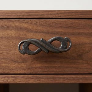 May include: Close-up of a dark bronze drawer pull with an infinity symbol design. The drawer pull is mounted on a wooden drawer with a natural wood grain finish. The hardware has a rustic, vintage aesthetic.