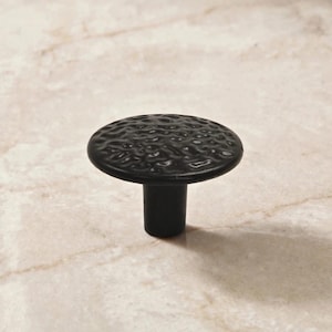 May include: A matte black cabinet knob featuring a circular top with a textured, uneven surface and a smooth, cylindrical base. The hardware has a dark, solid finish and a distinct, rippled pattern across the top face of the knob.