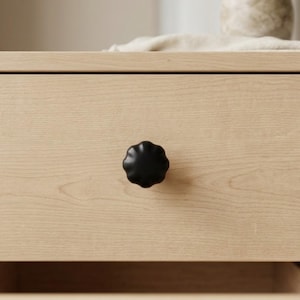May include: A close-up of a light-colored wooden drawer with a black, flower-shaped knob. The knob is centered on the drawer front, providing a striking contrast to the wood grain. The design suggests a modern or minimalist aesthetic.