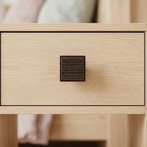 May include: Close-up of a light-colored wooden drawer with a dark brown square handle. The handle has a ridged design. The drawer is part of a larger piece of furniture, possibly a nightstand or dresser.