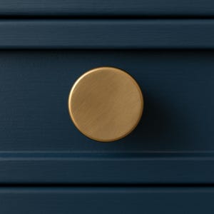 May include: A close-up of a gold-colored, round cabinet knob against a dark blue painted surface. The knob has a brushed metal finish and is centered in the frame. The background is a solid, dark blue color.