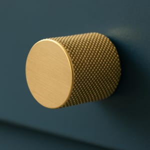 May include: A gold-colored cabinet knob with a cylindrical shape. The knob has a flat, circular face and a textured, dotted surface on the cylindrical body. The knob is mounted on a dark blue surface.