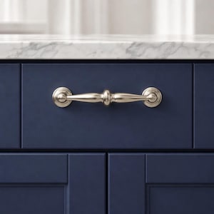 May include: A brushed nickel cabinet pull is centered on a navy blue cabinet door. The pull has a cylindrical center and rounded ends. The cabinet is part of a kitchen or bathroom setup, with a marble countertop visible above.