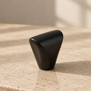 May include: A matte black cabinet knob with a distinct triangular, tapered shape. The knob features a smooth, solid surface with rounded edges and a wide, flat top that narrows toward the base, creating a geometric and functional hardware piece.