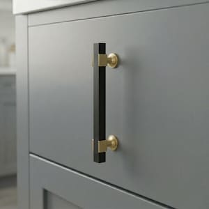 May include: A close-up of a gray cabinet door with a black and gold cabinet pull. The pull is rectangular with a black center and gold accents. The cabinet door is a solid gray color.