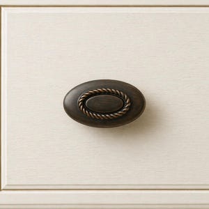 May include: Oval bronze cabinet pull with a rope-like border, mounted on a cream-colored wooden drawer front. The hardware has a dark, antique finish, providing a classic aesthetic. The drawer front has a subtle wood grain texture.
