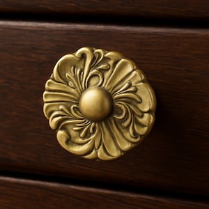 May include: Close-up of a gold-colored, ornate cabinet knob with a floral design. The knob has a central, rounded boss and is set against a dark brown wooden surface.