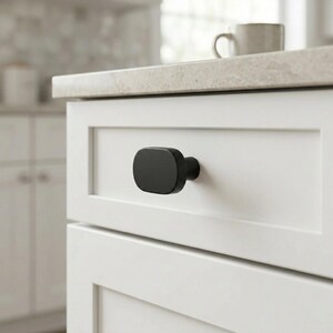 May include: A matte black cabinet knob with a rounded rectangular shape attached to a white shaker-style drawer front. The hardware features a smooth, solid finish and a central mounting point, providing a clean and functional accent for cabinetry.